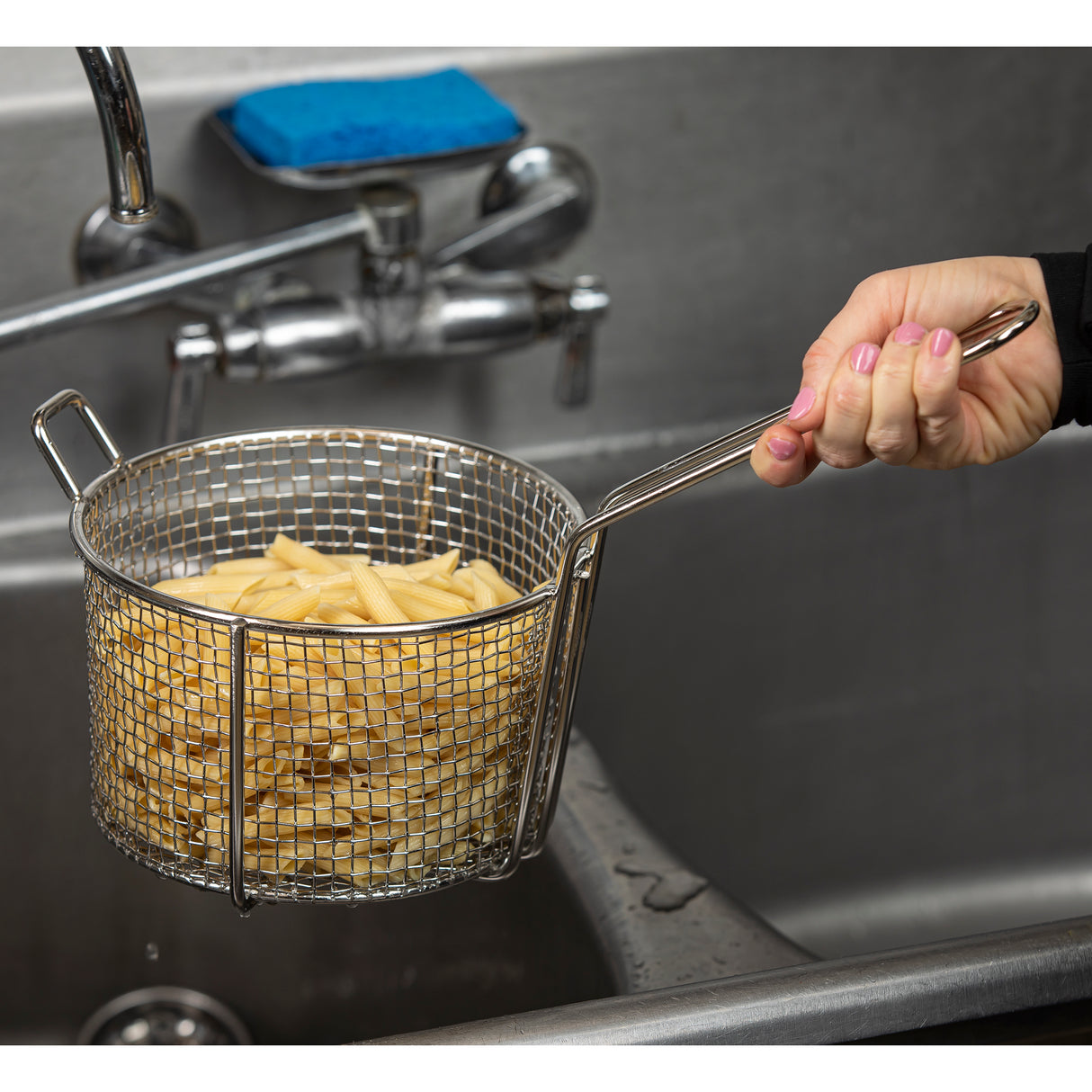 A hand holds a stainless steel cooking basket filled with cooked penne pasta over a sink. The faucet and a blue sponge rest nearby, and water droplets are visible, suggesting the pasta was just rinsed or drained.