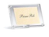 A clear acrylic sign holder displays a cream-colored card with white dots and elegant black script reading Prime Rib. The sign holder stands at a slight angle against a plain white background.