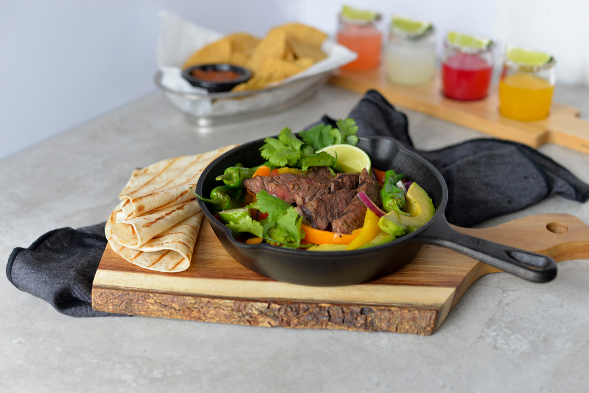 A black skillet with grilled steak, bell peppers, onions, and cilantro sits on a wood bread board with folded tortillas on the side. In the background, tortilla chips with salsa and three colorful drinks with lime wedges are visible on a gray surface.