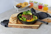 A black skillet with grilled steak, bell peppers, onions, and cilantro sits on a wood bread board with folded tortillas on the side. In the background, tortilla chips with salsa and three colorful drinks with lime wedges are visible on a gray surface.