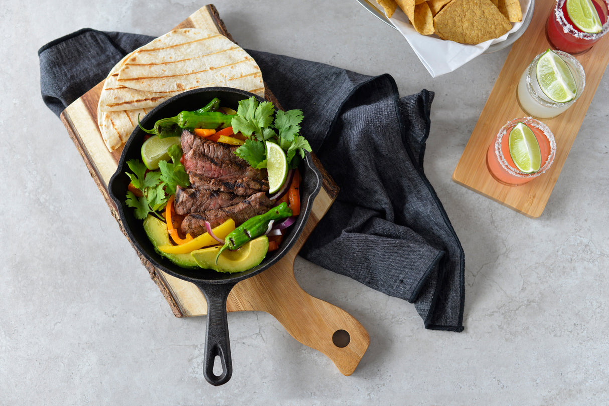 A cast-iron skillet with grilled steak strips, bell peppers, and green chili peppers sits on an Acacia bread board with folded tortillas. Fresh cilantro and a lime wedge garnish the skillet, alongside tortilla chips and three cocktails with lime slices.