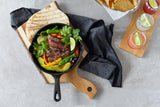 A cast-iron skillet with grilled steak strips, bell peppers, and green chili peppers sits on an Acacia bread board with folded tortillas. Fresh cilantro and a lime wedge garnish the skillet, alongside tortilla chips and three cocktails with lime slices.