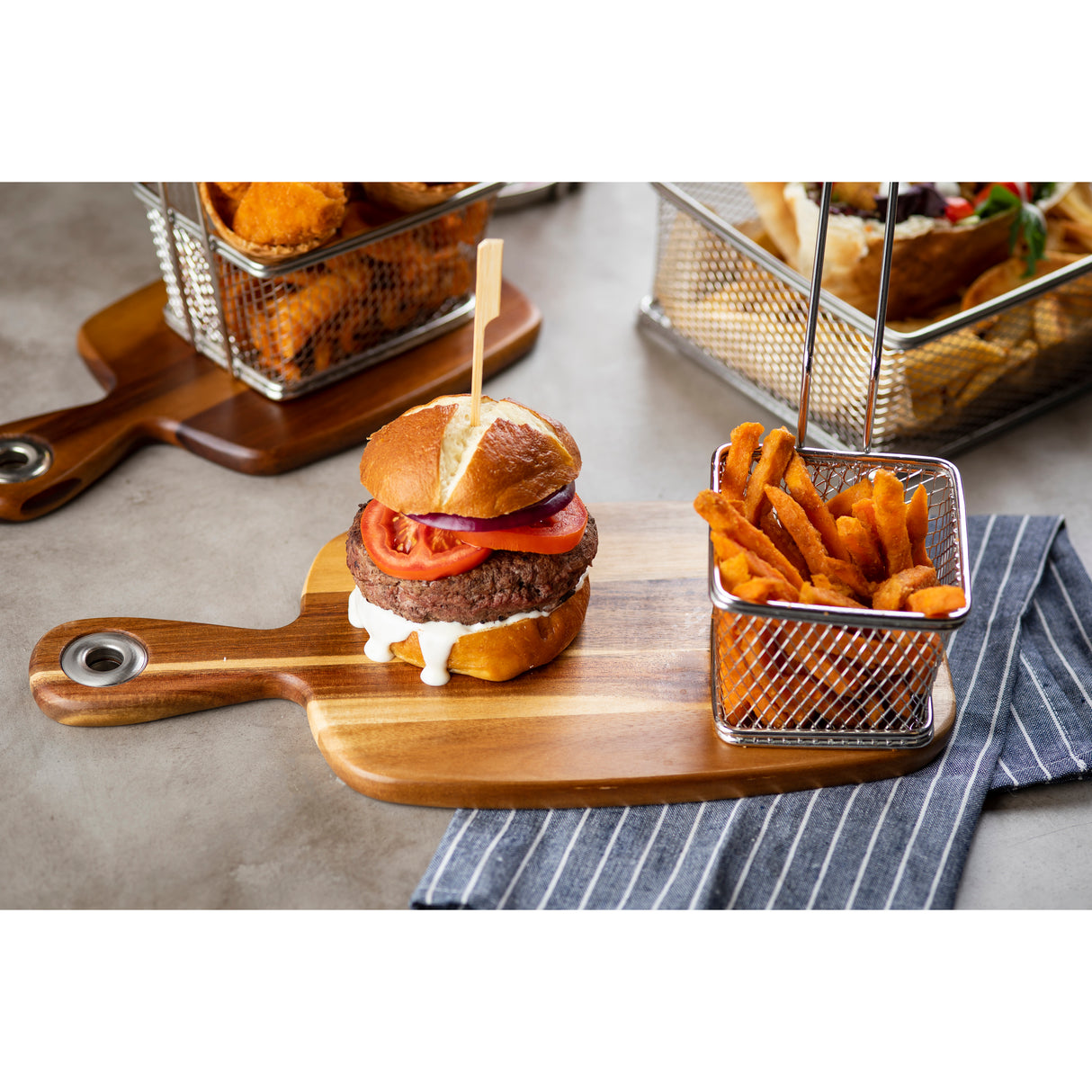 A juicy hamburger with tomato and lettuce rests on a TableCraft bread board beside a small metal basket of sweet potato fries. Other baskets of fried food sit in the background on a gray tabletop with a blue-and-white striped napkin nearby.