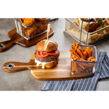 A juicy hamburger with tomato and lettuce rests on a TableCraft bread board beside a small metal basket of sweet potato fries. Other baskets of fried food sit in the background on a gray tabletop with a blue-and-white striped napkin nearby.
