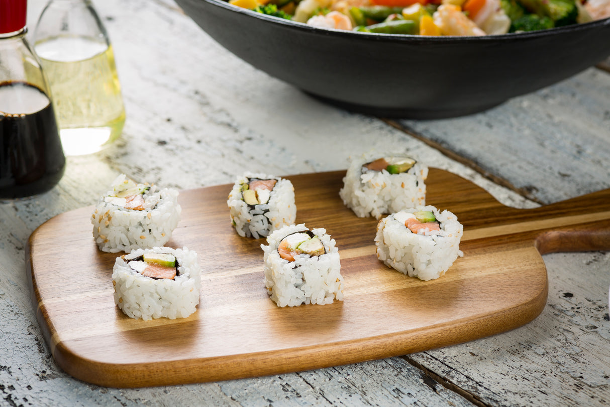 Six sushi rolls filled with avocado, cucumber, and fish are arranged on an acacia bread board. A bottle of soy sauce and a bowl of salad or stir-fry sit in the background, all on a rustic white wooden table.