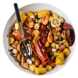 A large white bowl filled with a seafood boil featuring lobster, crab, shrimp, clams, corn on the cob pieces, red potatoes, and halved garlic bulbs. A colorful and rustic dish is served with a stainless steel slotted serving spoon on the side.