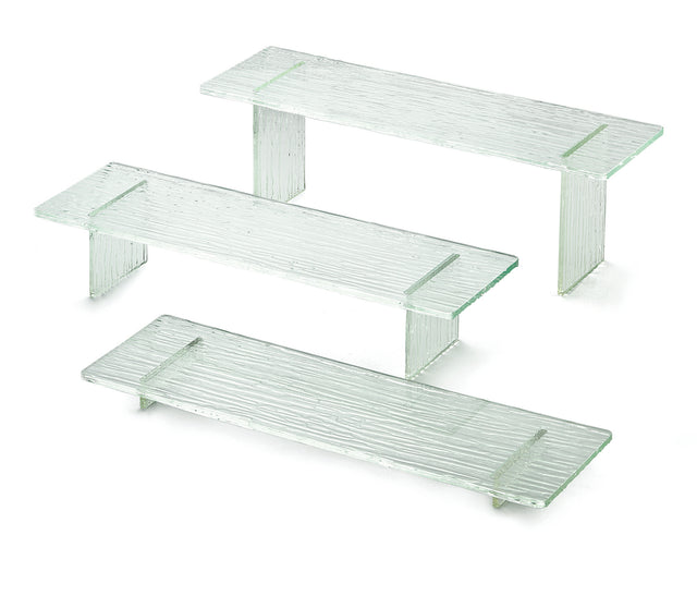 Three clear, textured glass shelves from the Cristal Collection Riser Set are displayed on a white background. Each shelf has two short side supports, creating minimalist, rectangular stands with a subtle striped pattern and transparent green accents.