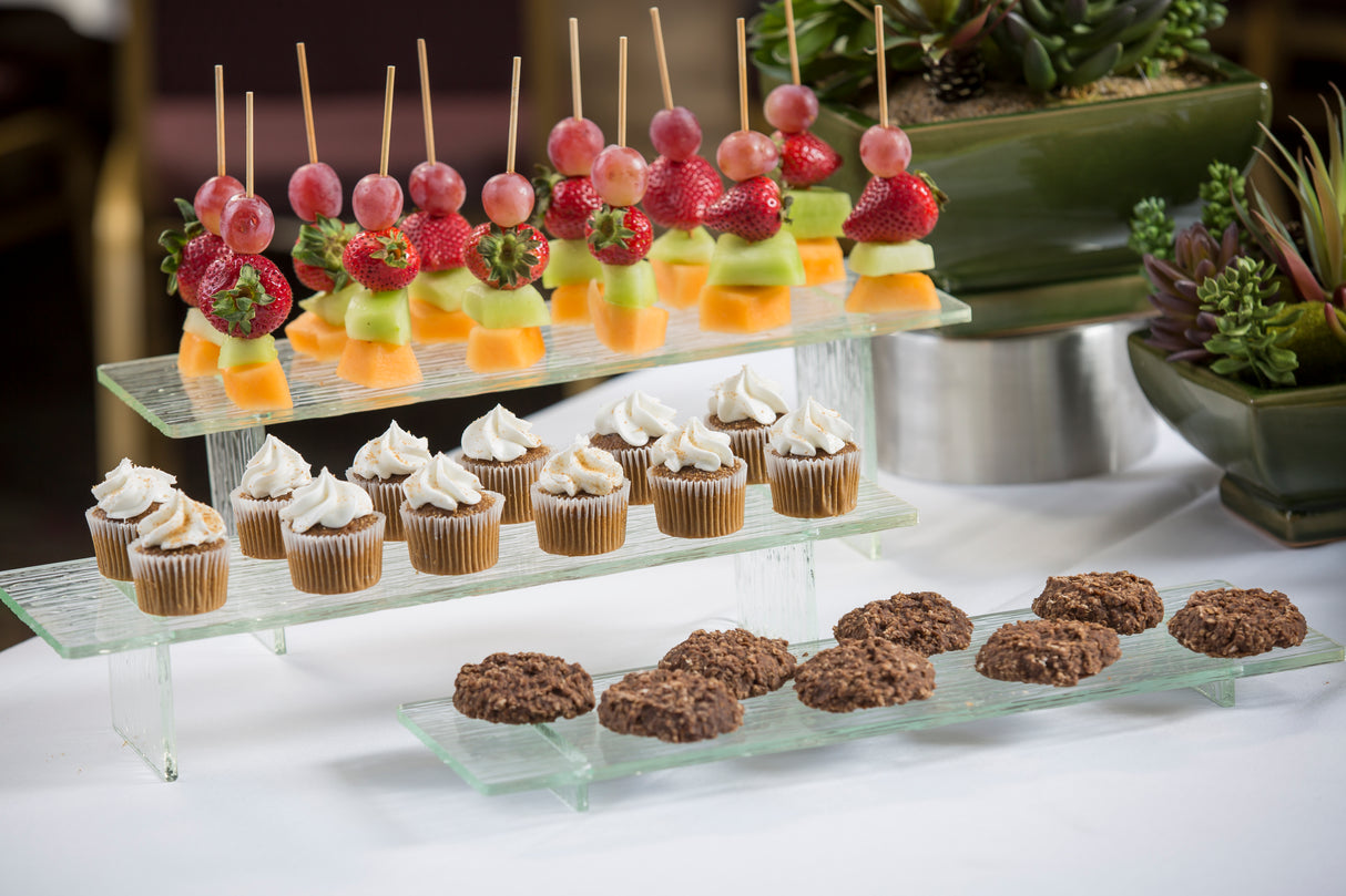 Three glass shelves, including a transparent green Cristal Collection Riser Set, display colorful fruit skewers on top, mini cupcakes in the middle, and round chocolate cookies below; lush plants decorate the background.