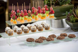 Three glass shelves, including a transparent green Cristal Collection Riser Set, display colorful fruit skewers on top, mini cupcakes in the middle, and round chocolate cookies below; lush plants decorate the background.
