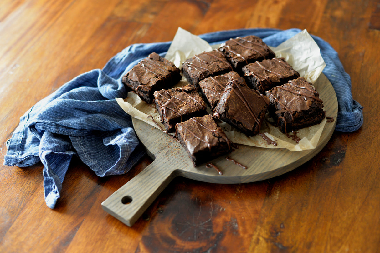 Nine chocolate brownies with a glossy drizzle are arranged on a TableCraft Ashwood paddle display board lined with parchment paper. The board sits on a wooden table next to a crumpled blue cloth, creating a rustic and inviting presentation.