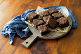Nine chocolate brownies with a glossy drizzle are arranged on a TableCraft Ashwood paddle display board lined with parchment paper. The board sits on a wooden table next to a crumpled blue cloth, creating a rustic and inviting presentation.