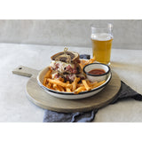 A plate with a stacked pastrami sandwich on rye, French fries, and ketchup sits on a TableCraft paddle board. Behind it is a glass of beer. The setup rests on a gray napkin against a light background.