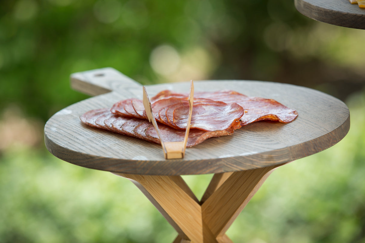 Thin slices of cured meat are arranged on a round Ashwood paddle board with a handle. A pair of wooden tongs rests on top. The serving board sits outdoors on a stand, framed by blurred greenery in the background.