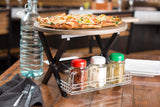A pizza on an Ashwood paddle board sits atop a black metal stand. Below, a rack holds four spice jars. Nearby are a glass water bottle, napkin, and utensils on a rustic wooden table in a restaurant setting.