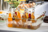 An acrylic beverage tub filled with ice holds various bottled soft drinks in vibrant colors. The dishwasher safe container sits on a table covered with a white tablecloth, with blurred food trays and bread loaves in the background.