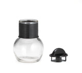 A clear, round glass Hottle with a wide base and a ribbed black plastic neck is shown. Next to it lies a matching lid with a grip knob on a white background. Both the Hottle and lid are dishwasher safe for easy cleaning.