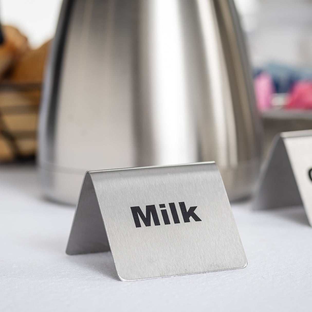 A small buffet tent metal milk sign with bold black letters sits on a white surface. In the blurred background, a stainless steel container and other items hint at a beverage or breakfast station.