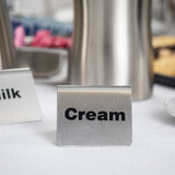 A TableCraft buffet tent in stainless steel labeled Cream in bold black letters sits on a white tablecloth. Part of another sign reading Milk and blurred containers appear in the background, suggesting a beverage or coffee station setup.