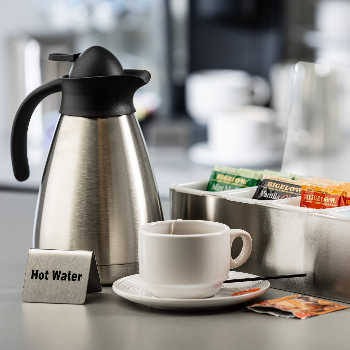 A stainless steel hot water carafe sits beside a white cup and saucer with a stirring stick, and a tray of assorted Bigelow tea bags. A small metal sign reads Hot Water. The softly blurred background hints at a buffet tent with additional cups.