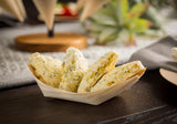 Four golden, oval-shaped scones speckled with herbs sit in a biodegradable pinewood boat shaped serving cup on a dark wooden table. The softly blurred background features a gray napkin and decorative green plants, creating a warm and inviting scene.
