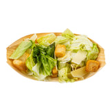 A small biodegradable pinewood boat serves as an eco-friendly disposable serving piece, holding a Caesar salad with romaine lettuce, golden croutons, and Parmesan shavings, all neatly arranged on a white background.