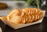 A TableCraft BAMDB5 biodegradable pinewood boat holds a neat row of golden-brown palmiers, crisp and flaky French pastries with a swirled, heart shape. Their caramelized surfaces shine atop an eco-friendly disposable serving piece.