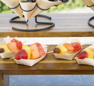 Small biodegradable pinewood boats hold colorful fruit skewers with strawberries, pineapple, watermelon, cantaloupe, and grapes. Arranged on a wooden shelf, these eco-friendly disposable serving pieces create a fresh and summery scene.