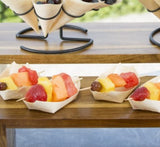 Small biodegradable pinewood boats hold colorful fruit skewers with strawberries, pineapple, watermelon, cantaloupe, and grapes. Arranged on a wooden shelf, these eco-friendly disposable serving pieces create a fresh and summery scene.
