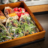 A wooden tray holds packaged salads with spinach, pecans, dried cranberries, and cheese, wrapped in plastic, alongside cheese wheels and small pinewood boat shaped biodegradable serving pieces with sliced meats and cheese. The tray sits on a dark surface by a fridge.