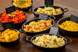 A variety of macaroni and cheese dishes in black serving bowls, each labeled with a TableCraft chalkboard bamboo pick: Flamin’ Hot Mac, Bacon Mac, Cheesy Wheels, Maine Lobster Mac & Cheese, White Cheddar Macaroni, and Creamy Cheddar Mac on a wooden table.