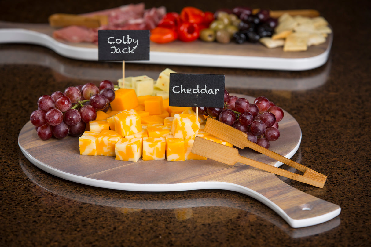 A wooden serving board with cubed Colby Jack and Cheddar cheeses, labeled with TableCraft chalkboard signs, is flanked by red grapes and wooden tongs. In the background, another board holds crackers, olives, red peppers, and sliced meats.