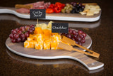A wooden serving board with cubed Colby Jack and Cheddar cheeses, labeled with TableCraft chalkboard signs, is flanked by red grapes and wooden tongs. In the background, another board holds crackers, olives, red peppers, and sliced meats.