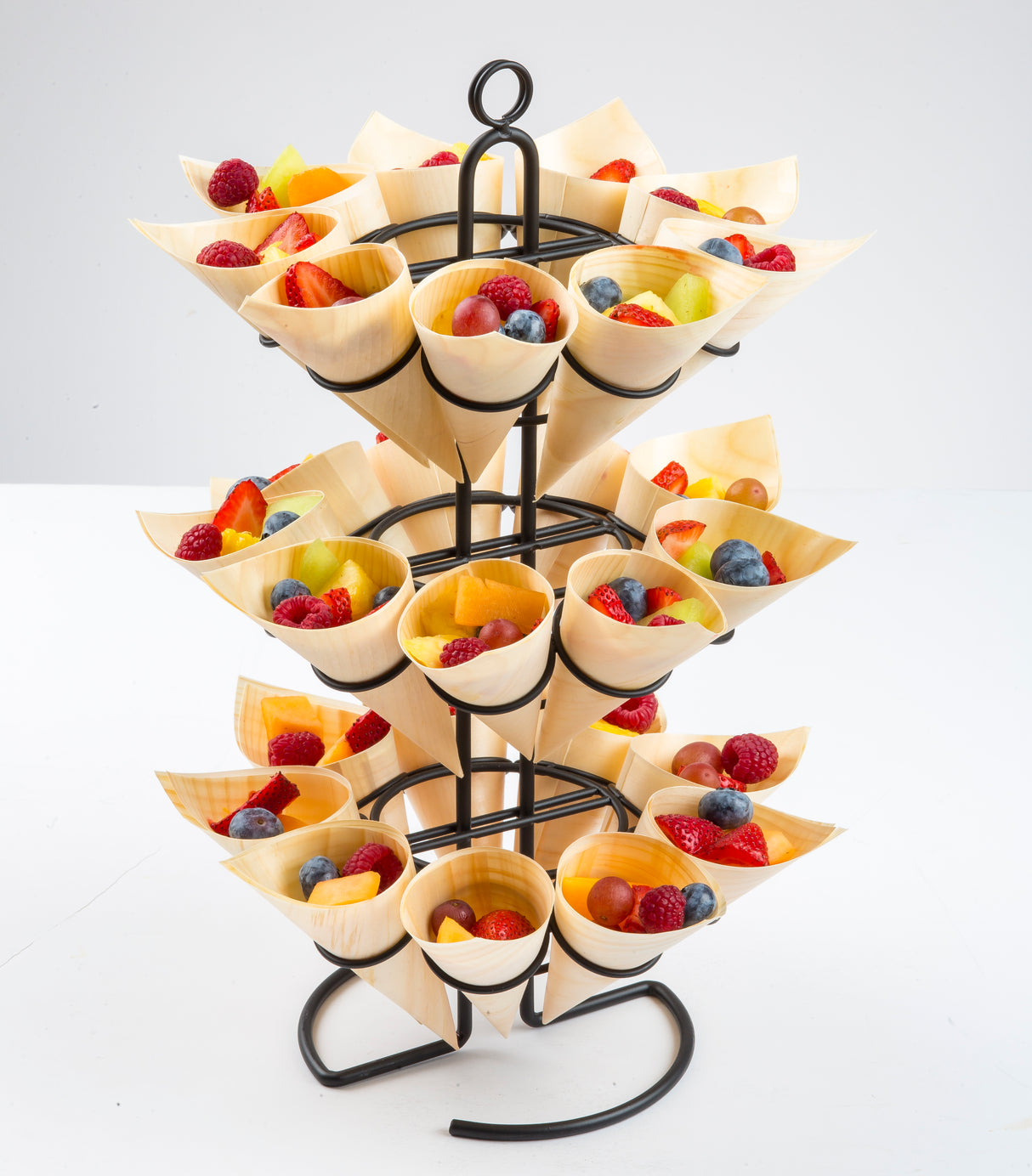A black metal stand holds multiple eco-friendly, biodegradable disposable serving cones made of light-colored wood, each filled with fresh fruits like strawberries, blueberries, raspberries, grapes, and kiwi against a plain white background.
