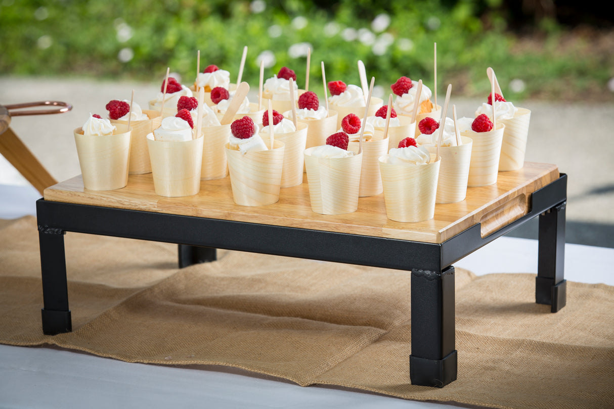 A wooden tray on black metal legs holds small, eco-friendly disposable serving cups of whipped cream topped with red raspberries, each with a wooden spoon. Arranged in rows, the tray sits on a burlap runner outdoors against blurred greenery.