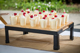 A wooden tray on black metal legs holds small, eco-friendly disposable serving cups of whipped cream topped with red raspberries, each with a wooden spoon. Arranged in rows, the tray sits on a burlap runner outdoors against blurred greenery.