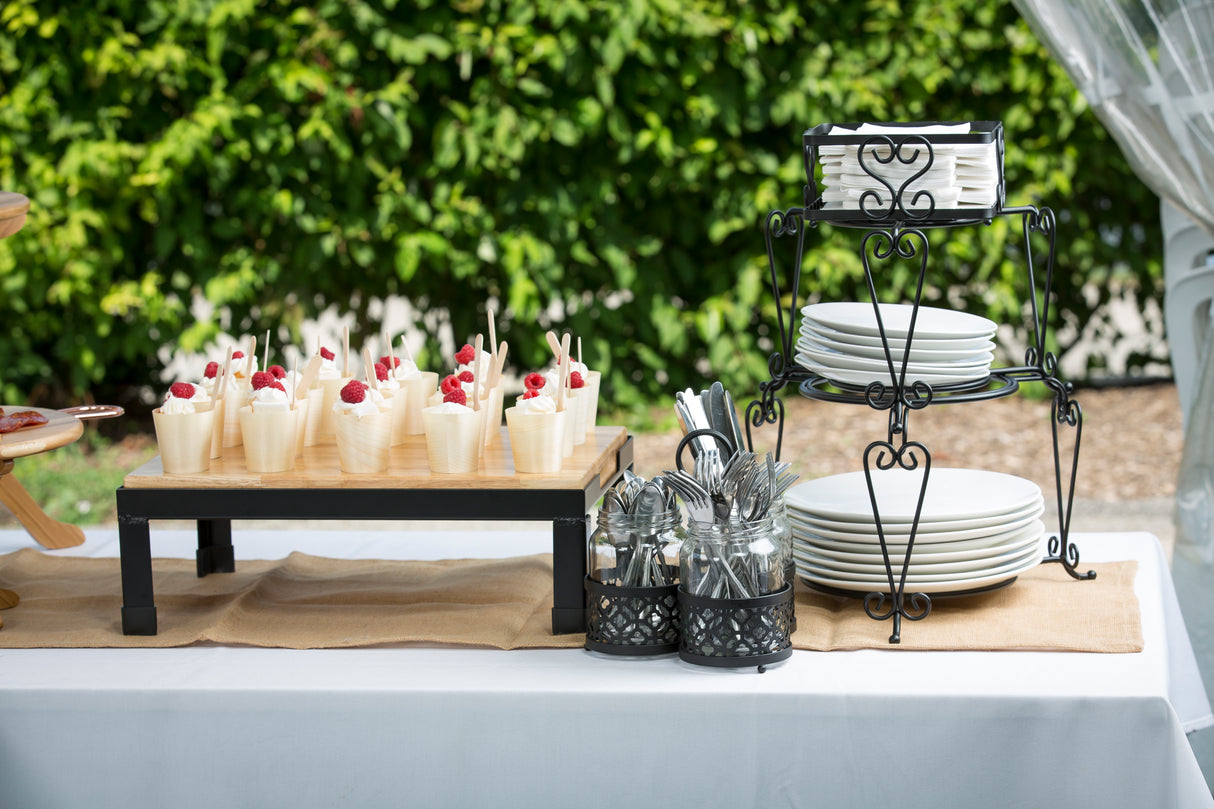 A buffet table outdoors displays desserts in biodegradable, disposable serving cups topped with raspberries, a stand holding stacked white plates and napkins, and containers of silver utensils, all arranged on a white tablecloth with a tan runner amid green foliage.