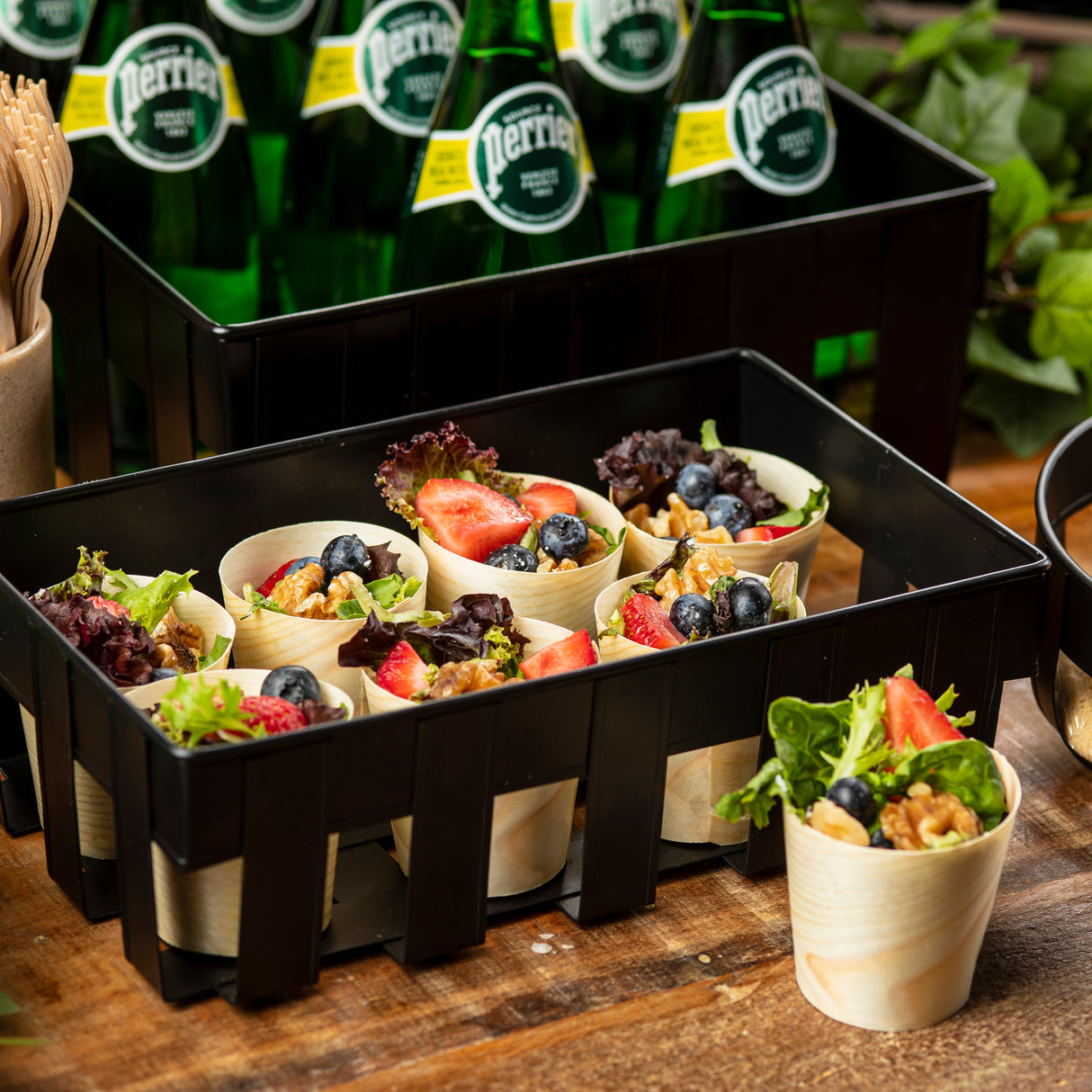 A black crate holds eco-friendly, biodegradable paper cups filled with mixed greens, walnuts, strawberries, and blueberries. Perrier bottles are stacked behind, wooden cutlery is visible to the side, and green leaves add a rustic touch.