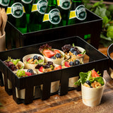 A black crate holds eco-friendly, biodegradable paper cups filled with mixed greens, walnuts, strawberries, and blueberries. Perrier bottles are stacked behind, wooden cutlery is visible to the side, and green leaves add a rustic touch.