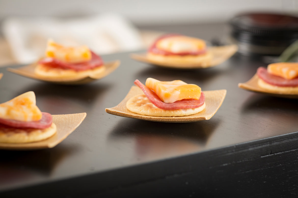 Close-up of crackers topped with salami and marbled cheddar cheese, each served on a small bamboo plate, displayed on a dark glossy surface. The softly blurred background draws attention to the delicious appetizers in the foreground.
