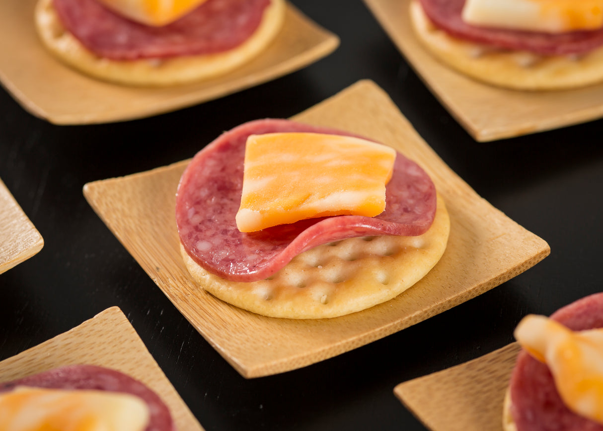 A square bamboo disposable dish holds a round cracker topped with salami and marbled cheddar cheese. Other similar TableCraft plates are partially visible on a black surface in the background.