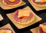 A square bamboo disposable dish holds a round cracker topped with salami and marbled cheddar cheese. Other similar TableCraft plates are partially visible on a black surface in the background.