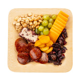 A square bamboo plate holds marcona almonds, green grapes, cheddar cheese slices, dried apricots, dried cranberries, pepperoni slices, and a small wedge of cranberry cheese, all arranged in neat sections.
