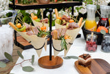 A table set outdoors displays paper cones filled with salami, cheese, breadsticks, kiwi, and cherry tomatoes, each artfully garnished with rosemary and secured by a bamboo knot pick from TableCraft. Lush plants surround jars of juice and berries and a bucket of ice.
