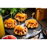 Four mini waffles topped with fried chicken are each secured with a bamboo knot pick, served on a black tray. Green leaves, orange juice, and strawberries add to the bright and inviting setting in the background.