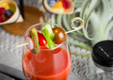 A glass of red tomato juice garnished with a bamboo Card Holder Pick, skewering a cherry tomato, cucumber, red pepper, radish slice, and a small pickle. Blurred appetizers and a green plant create a vibrant, fresh setting.