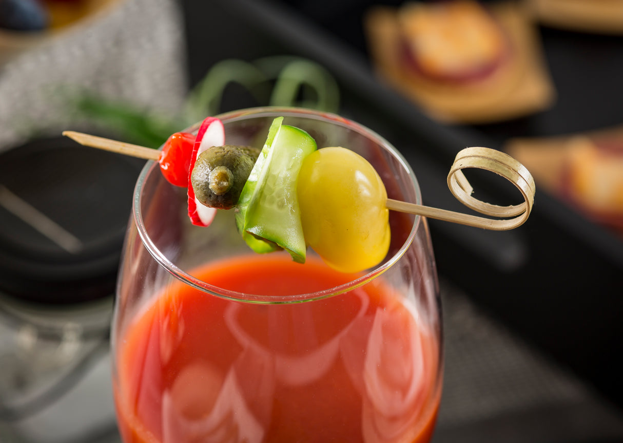 A cocktail glass filled with a red drink, topped with a bamboo Card Holder Pick holding a yellow cherry tomato, cucumber ribbon, caperberry, and red radish slice. The background features a blurred tray with more appetizers.