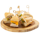 Five sandwich quarters made with baguette, turkey, cheddar cheese, and lettuce are arranged on a round wooden serving board. Each is secured with a decorative TableCraft bamboo card holder pick. The background is plain white.