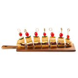 A wooden serving board holds six mini sandwich bites, each skewered with a bamboo card holder pick and topped with a cherry tomato and cucumber slice. The sandwiches, arranged neatly in a row, feature baguette bread, cheese, and a layer of dark filling.