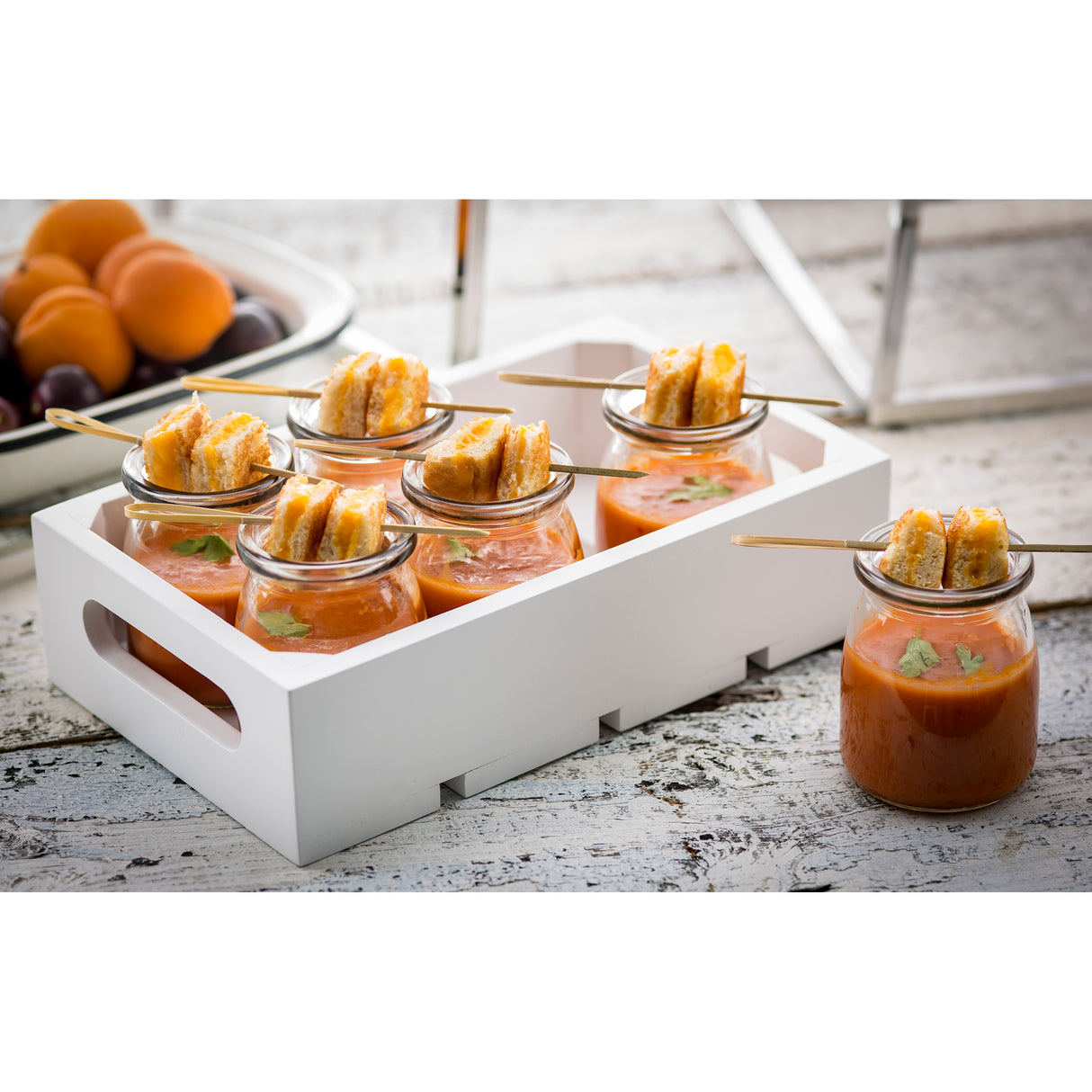 Six small glass jars of tomato soup, each topped with mini grilled cheese sandwiches skewered on a bamboo pick, are arranged in a white wooden tray on a rustic white surface. A bowl of apricots and plums sits in the background.
