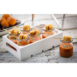 Six small glass jars of tomato soup, each topped with mini grilled cheese sandwiches skewered on a bamboo pick, are arranged in a white wooden tray on a rustic white surface. A bowl of apricots and plums sits in the background.